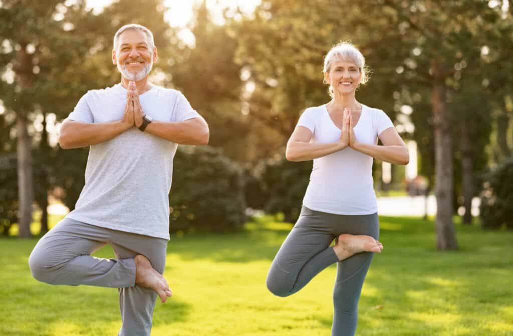 A senior man and a senior woman smiling and doing yoga poses outside.
